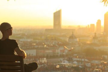 A bald man wearing glasses and relaxing on a park bench overlooking a sprawling city at sunrise.