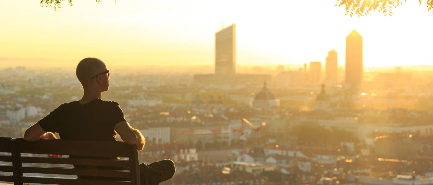 A bald man wearing glasses and relaxing on a park bench overlooking a sprawling city at sunrise.