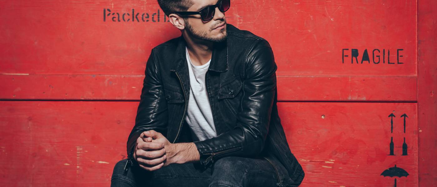 A stylish young man wearing a white t-shirt under a black leather jacket, sitting on a wooden crate in front of a red wall.