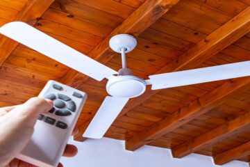 A man's hand reaches out with a remote to adjust his ceiling fan, which spins on a ceiling made of brown wooden beams.