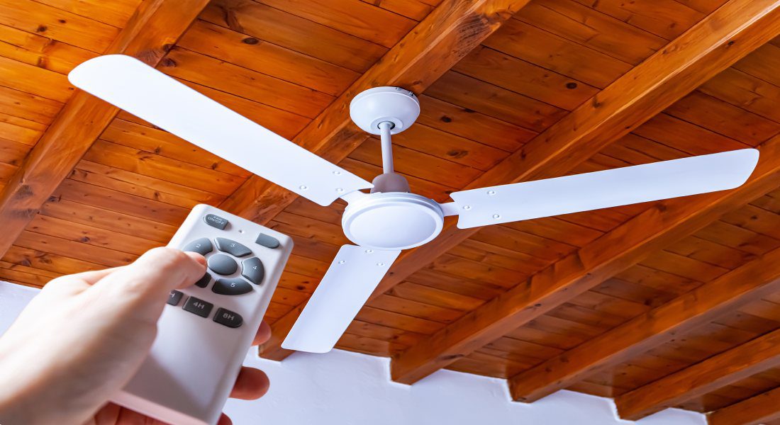 A man's hand reaches out with a remote to adjust his ceiling fan, which spins on a ceiling made of brown wooden beams.