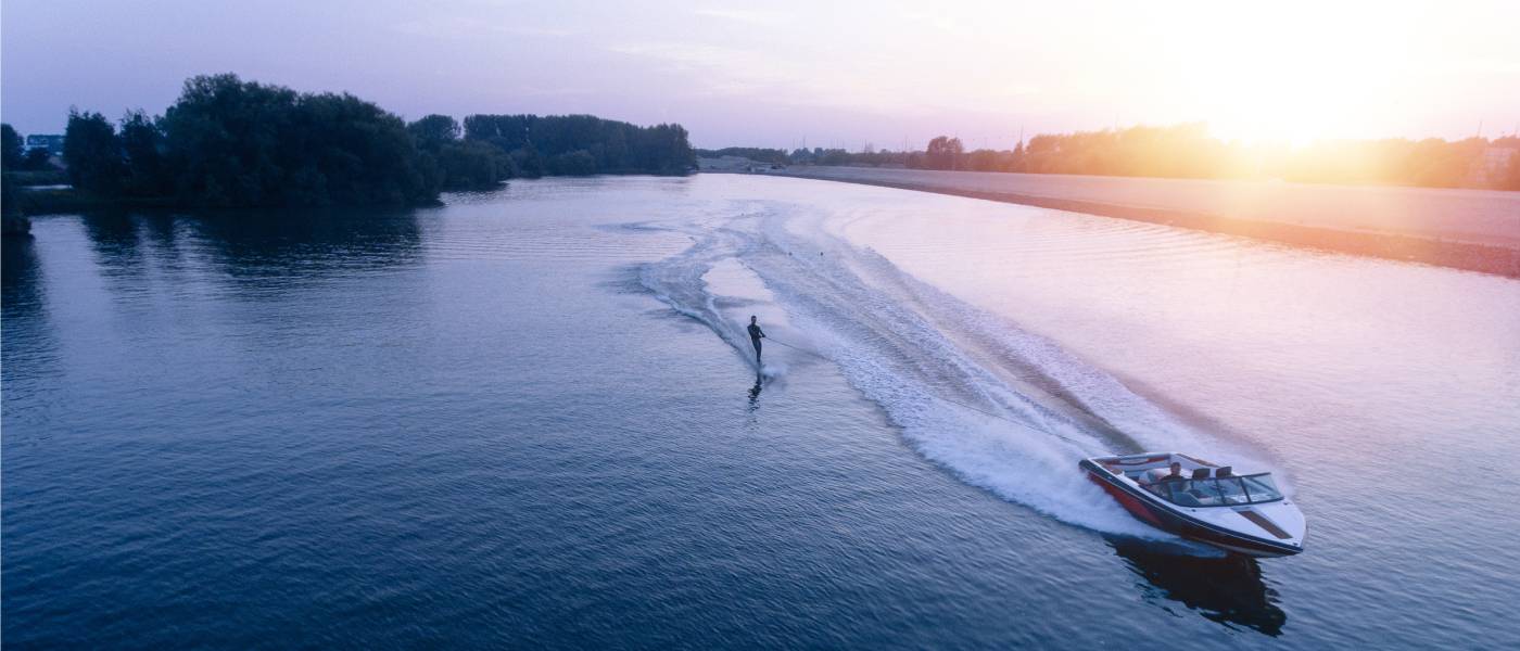 A pair of friends boating on the lake with one driving the speed boat and another trailing behind on a ski.