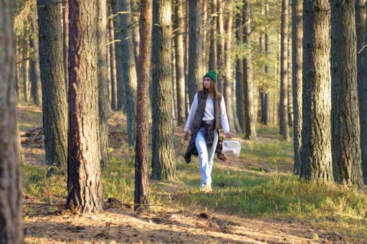 A woman wearing a green hat with a flannel shirt tied around her waist walks through the woods with a basket.