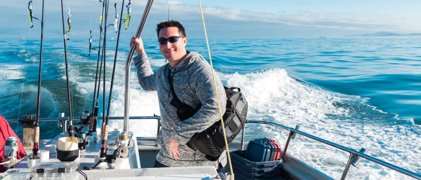 A happy angler on a fishing boat, smiling, as he's surrounded by several fishing poles. The boat is moving to a fishing location.