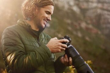 A man wearing outdoor gear smiling down at his professional camera. He’s standing in a grassy area with mountains behind him.