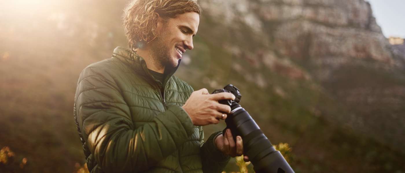 A man wearing outdoor gear smiling down at his professional camera. He’s standing in a grassy area with mountains behind him.