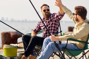 Two guys sitting in camping chairs and fishing on a beach. They're giving each other a big high five as their rods are cast out toward the water.