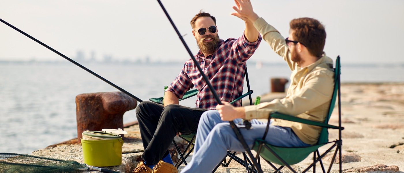 Two guys sitting in camping chairs and fishing on a beach. They're giving each other a big high five as their rods are cast out toward the water.