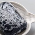 A foamy black bar of hand soap sitting in a wavy, nautical-themed ceramic soap dish on a plain surface.