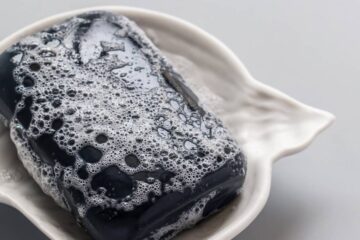 A foamy black bar of hand soap sitting in a wavy, nautical-themed ceramic soap dish on a plain surface.