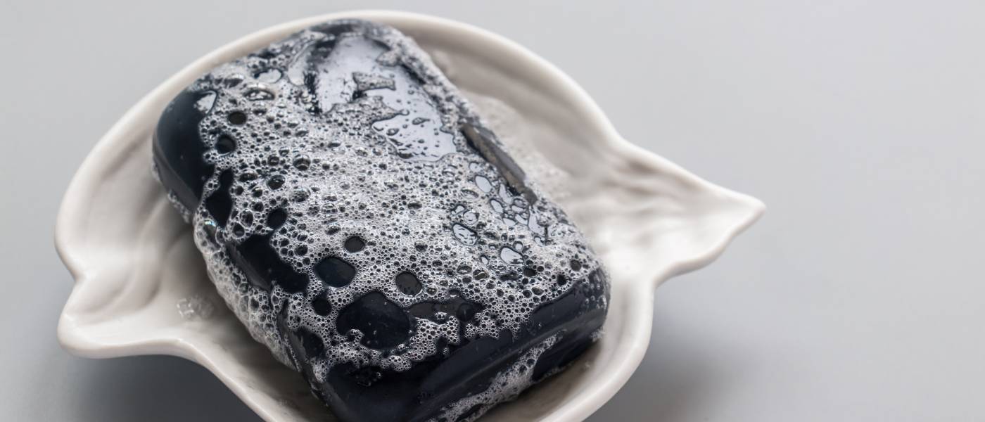 A foamy black bar of hand soap sitting in a wavy, nautical-themed ceramic soap dish on a plain surface.