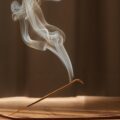 A flat incense burner on a hardwood floor with a lit stick of incense placed in it. The smoke is creating interesting swirls.