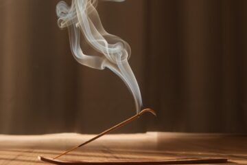 A flat incense burner on a hardwood floor with a lit stick of incense placed in it. The smoke is creating interesting swirls.