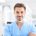 A young male dentist in scrubs smiling at the camera in an examining room. A disposable face mask is secured under his chin.