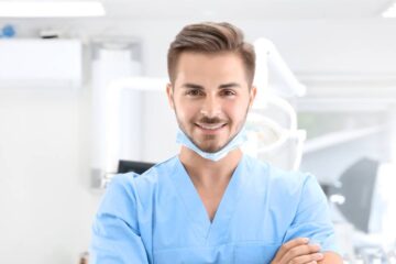 A young male dentist in scrubs smiling at the camera in an examining room. A disposable face mask is secured under his chin.