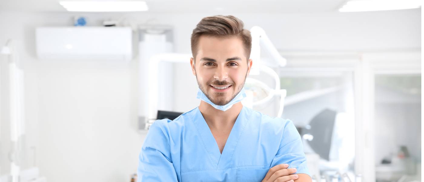 A young male dentist in scrubs smiling at the camera in an examining room. A disposable face mask is secured under his chin.