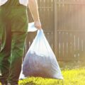 A person wearing green overalls carrying two bags of trash in a yard. The lawn is overgrown with sunlight shining on it.