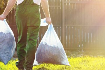 A person wearing green overalls carrying two bags of trash in a yard. The lawn is overgrown with sunlight shining on it.