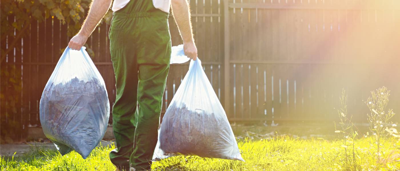 A person wearing green overalls carrying two bags of trash in a yard. The lawn is overgrown with sunlight shining on it.