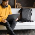 A man wearing a yellow jumper sits on a grey couch in a hotel room on his laptop next to his yellow luggage.