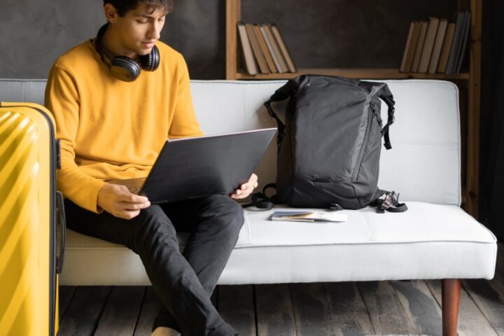 A man wearing a yellow jumper sits on a grey couch in a hotel room on his laptop next to his yellow luggage.