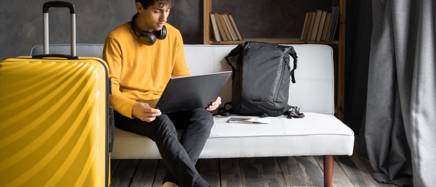 A man wearing a yellow jumper sits on a grey couch in a hotel room on his laptop next to his yellow luggage.