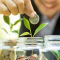 A blurred young man dropping a coin into the middle of three glass jars in a row, each filled with coins and sprouts.