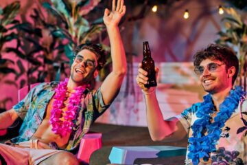 Two men partying together at a house party. They're both sitting down, laughing, and drinking out of a glass bottle.