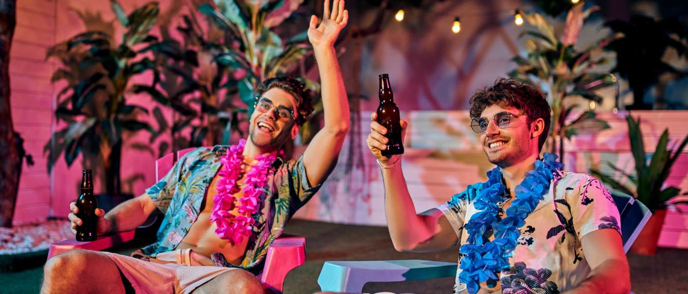 Two men partying together at a house party. They're both sitting down, laughing, and drinking out of a glass bottle.