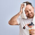 A man in casual athletic wear leaning on a wall and wiping sweat from his forehead while staring down at his protein shake.