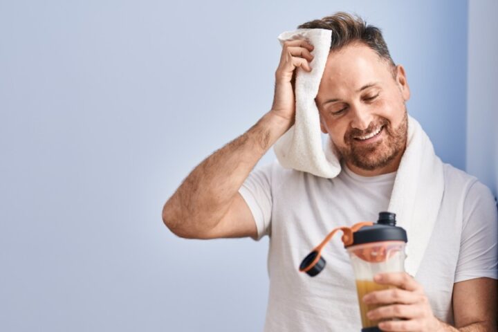 A man in casual athletic wear leaning on a wall and wiping sweat from his forehead while staring down at his protein shake.