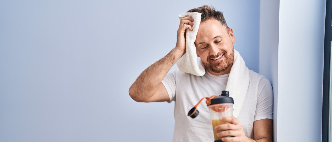 A man in casual athletic wear leaning on a wall and wiping sweat from his forehead while staring down at his protein shake.