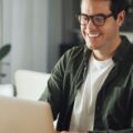 A young man wearing glasses and smiling as he sits at a desk in a home and types on his silver laptop.