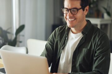 A young man wearing glasses and smiling as he sits at a desk in a home and types on his silver laptop.