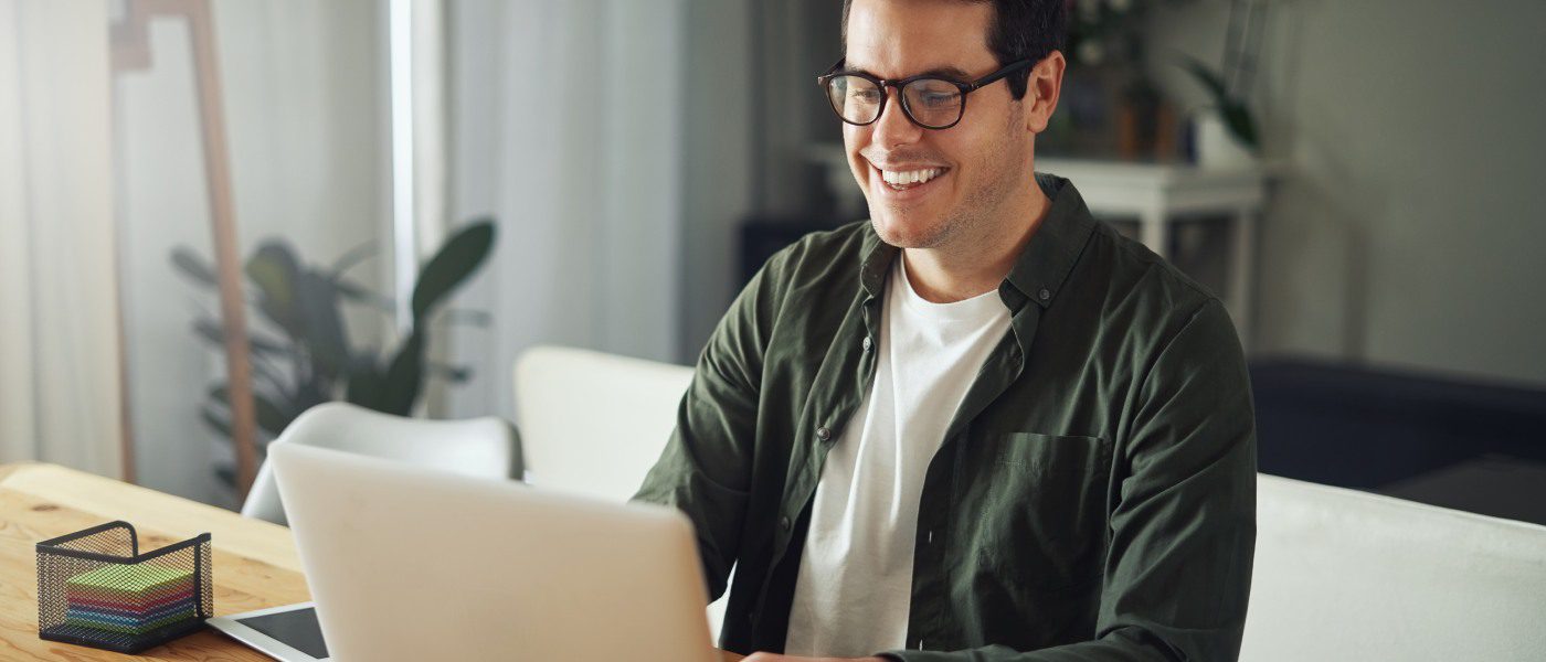 A young man wearing glasses and smiling as he sits at a desk in a home and types on his silver laptop.