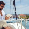 A young man wearing sitting on the deck of a yacht while reading a tablet on a sunny day. A coffee mug is beside him.