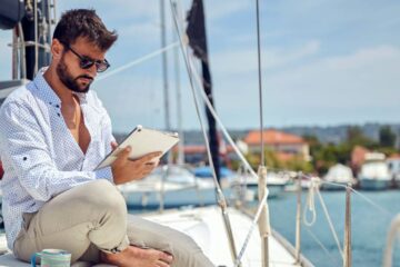 A young man wearing sitting on the deck of a yacht while reading a tablet on a sunny day. A coffee mug is beside him.