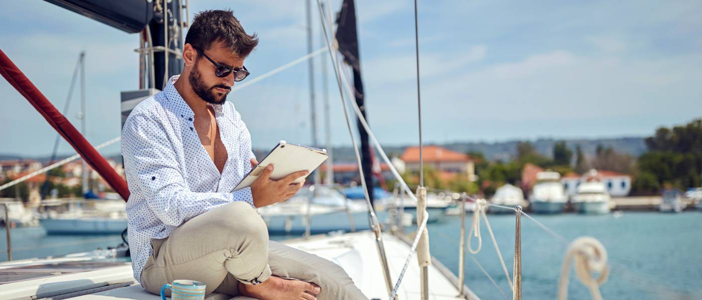 A young man wearing sitting on the deck of a yacht while reading a tablet on a sunny day. A coffee mug is beside him.