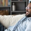 A relaxed man in nice yet casual attire lounging on a white couch in a stylish, well-organized room.