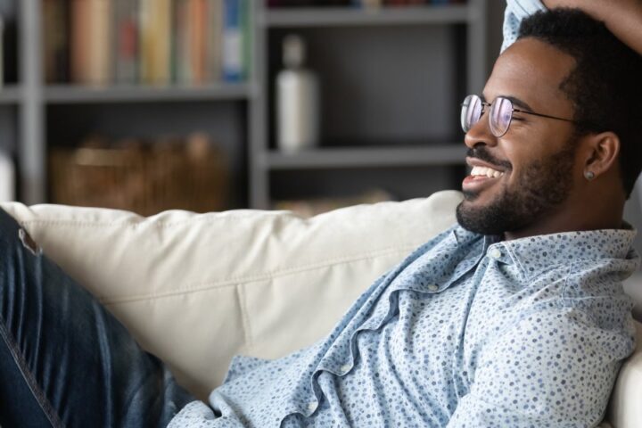 A relaxed man in nice yet casual attire lounging on a white couch in a stylish, well-organized room.