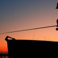 An outdoorperson standing in a boat enjoying a night fish. The sun is going down behind them and the line is in the water.