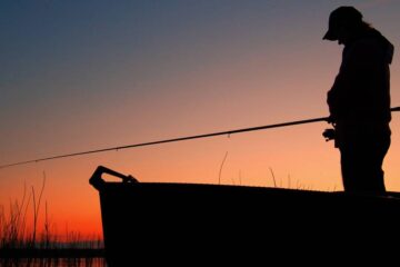 An outdoorperson standing in a boat enjoying a night fish. The sun is going down behind them and the line is in the water.