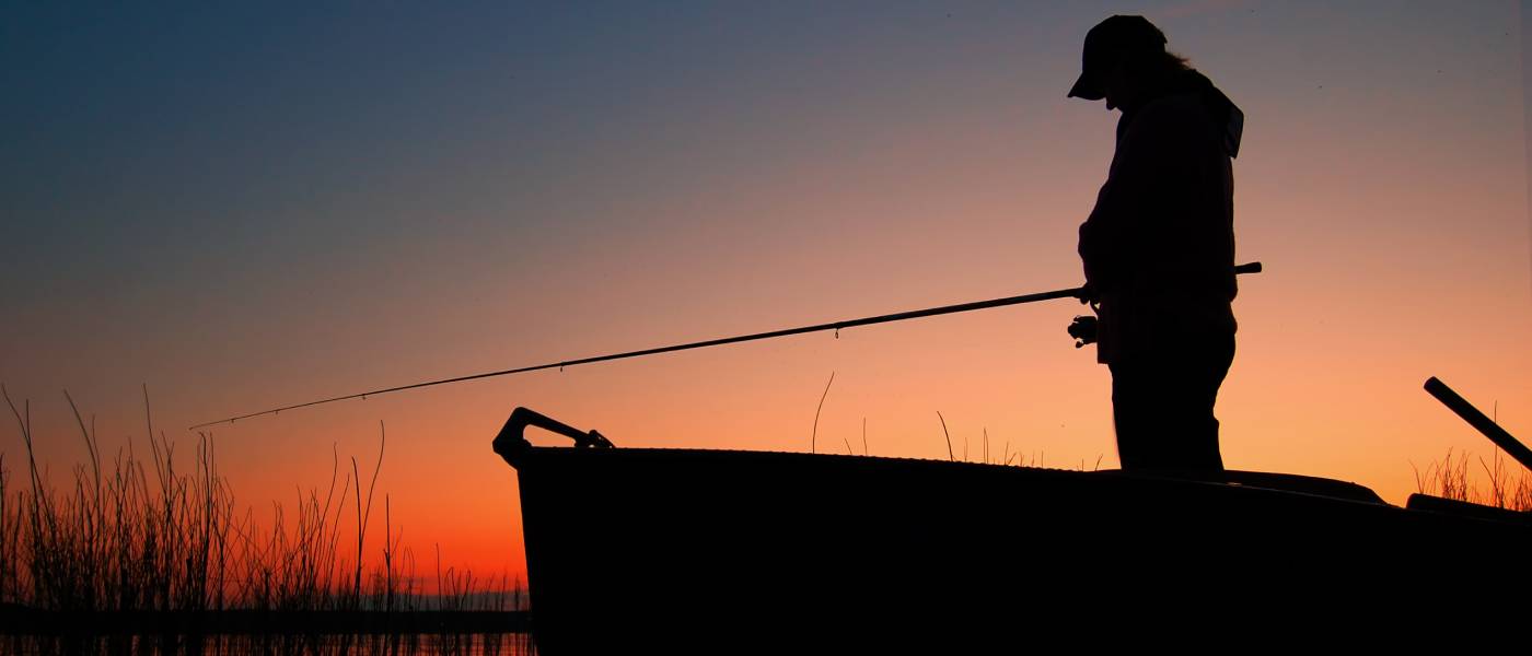 An outdoorperson standing in a boat enjoying a night fish. The sun is going down behind them and the line is in the water.