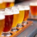 A close-up to a flight of craft beer with tall glasses filled with different colors of beer on top of a bench with a blurry background.