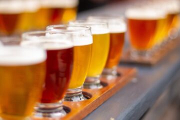 A close-up to a flight of craft beer with tall glasses filled with different colors of beer on top of a bench with a blurry background.