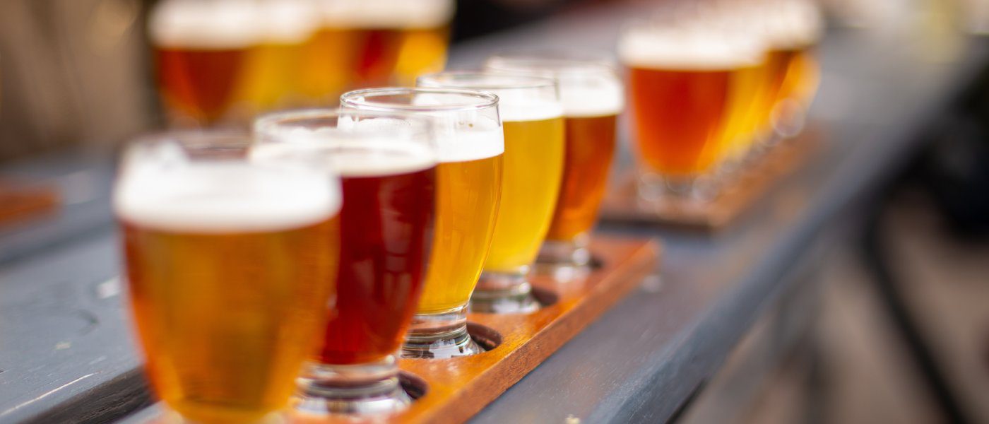 A close-up to a flight of craft beer with tall glasses filled with different colors of beer on top of a bench with a blurry background.