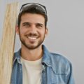 A young man in a denim jacket and white tee shirt smiles while holding a piece of plywood in a white room.