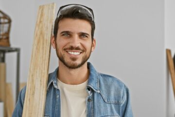 A young man in a denim jacket and white tee shirt smiles while holding a piece of plywood in a white room.