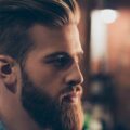 A handsome man standing with his side profile exposed. He has brown hair, nicely groomed and a grown out beard.