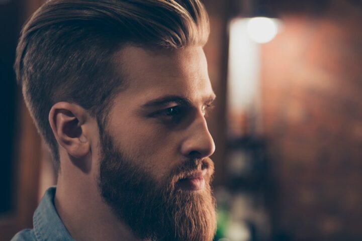 A handsome man standing with his side profile exposed. He has brown hair, nicely groomed and a grown out beard.